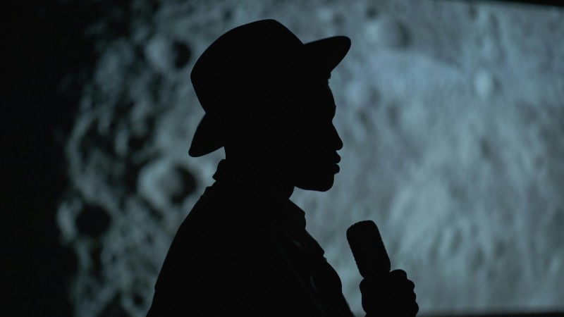 A dramatic silhouette of a person with a hat holding a microphone against a moon-like background., tags: poetry-film-festivals - pexel